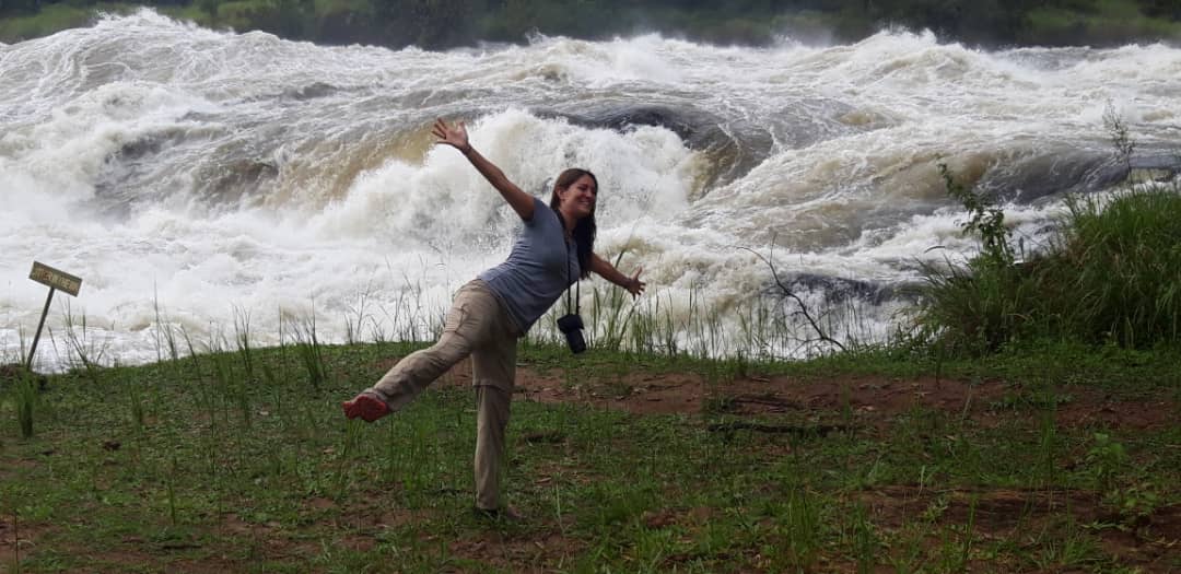 A joyful clients' poses with outstretched arms and one leg lifted in front of the powerful Murchison Falls, with turbulent white water cascading in the background. Murchison Falls National Park Uganda